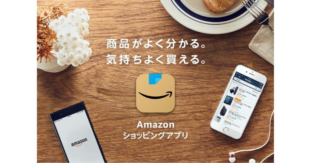 木目のテーブルの上に置かれた2台のスマートフォン。中央にはAmazonのアイコンと「Amazonショッピングアプリ」の文字、上部には「商品がよく分かる。気持ちよく買える。」というキャッチコピーが表示されている。