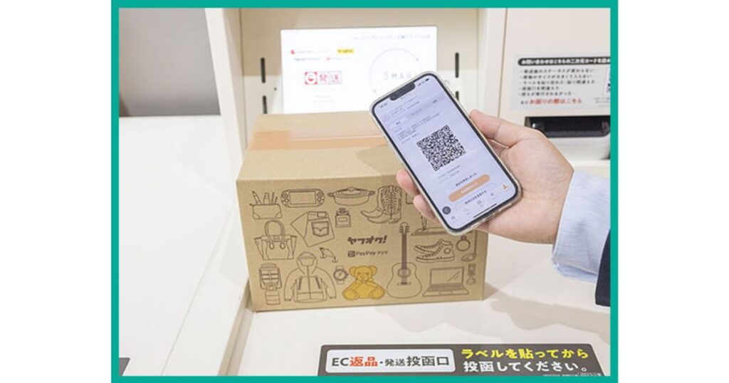ヤフオクやPayPayフリマのロゴがついたダンボール箱の横で、発送用バーコード画面を表示したスマートフォンを手に持っている実写画像。