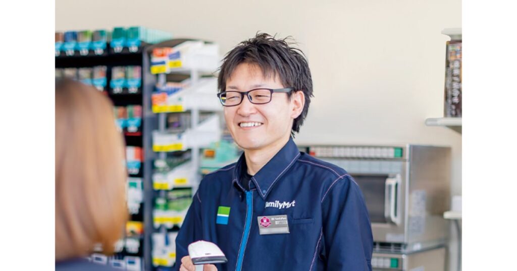 コンビニのレジカウンターで、笑顔で接客対応をしているメガネをかけた男性店員の写真。