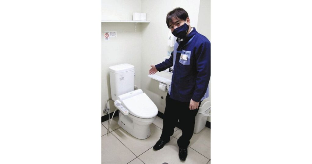 コンビニの個室トイレ内で、黒いマスクと制服を着用した男性店員が、洋式便器を案内するように手を差し向けている写真。