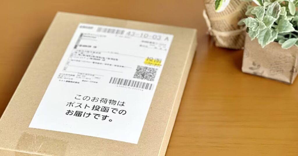 机の上に置かれた、送り状と「このお荷物はポスト投函でのお届けです。」という案内シールが貼られた薄型の発送用段ボール箱。